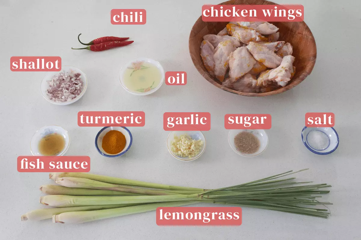 A bowl of chicken wings along with lemongrass and chili alongside dishes of chopped red shallots, oil, turmeric powder, fish sauce, finely chopped garlic, sugar and salt.