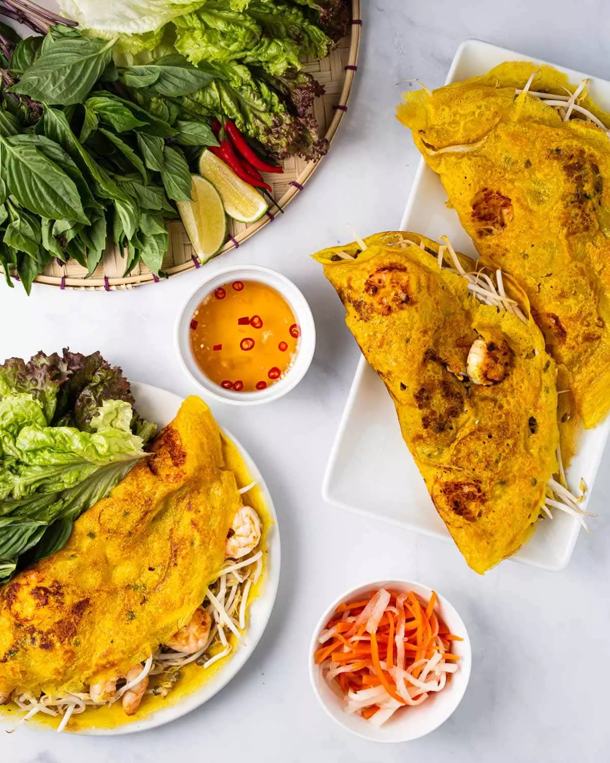 Plate of banh xeo surrounded by assorted greens, dipping sauce, serving tray of banh xeo, and pickled carrots and daikon.